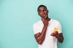 Young African American man holding a mobile phone isolated on blue background looking sideways with doubtful and skeptical expression.