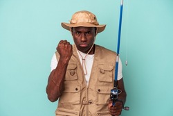 Young African American fisherman holding rod isolated on blue background showing fist to camera, aggressive facial expression.