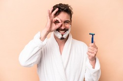 Young caucasian man wearing shaving foam and holding razor blade isolated on beige background excited keeping ok gesture on eye.