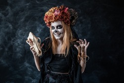 red-haired woman in Halloween costume, witch make-up with horns, dried flowers on her head conjures in dark smoky room, looks at camera, smiles maliciously, holding skull of wolf, clawed legs of eagle