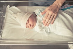 Mother checking on newborn baby boy sleeping in hospital crib.