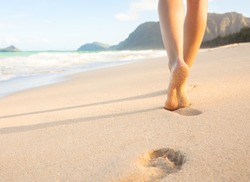 Beach travel - woman walking on sand beach leaving footprints in the sand. Closeup detail of female feet and golden sand on beach in Hawaii.
