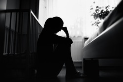 Silhouette of tired, stressed mother sitting next to baby crib at home. 