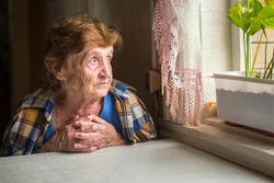 Old lonely woman sitting near the window in his house.