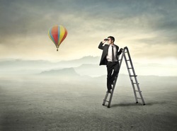 Young businessman on a ladder using binoculars and hot-air balloon in the background