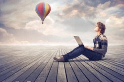 Young man sitting on a wooden floor and using a laptop with hot-air balloon in the background