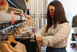 Beautiful young woman in shop looks at clothes to buy wearing protective face mask during Coronavirus pandemic Covid-19 - Shop assistant arranges dress