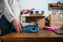 Business woman prepares a parcel for shipping in a cardboard box with clothes from her online store - Millennial sells second-hand used dresses in her home using the smartphone application - Start up