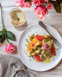 Gourmet white fish recipe with assorted citrus fruits, olives, fresh herbs, marinated red onion and fennel, served on a white ceramic plate and with a glass of white wine. Table decoration with roses