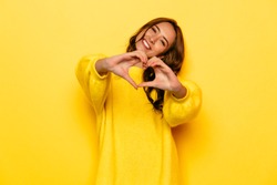 Smiling young girl in yellow sweater showing heart with two hands, love sign. Isolated over yellow background.