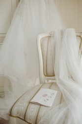 Wedding rings lie on a wedding invitation on a white beautiful chair in a bright room against the background of the bride's white dress