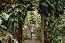 Young woman walking in tropical garden in long summer dress, greenery and palm trees around, enjoying nature