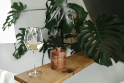 Gass of white wine on a wooden stand in a bathroom with tropical plants. Vacation home concept.