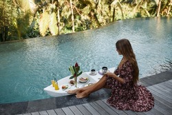 Beautiful slender girl in a red dress has breakfast sitting by the pool. Floating breakfast in tropical resort. Table relaxing in calm pool water, healthy breakfast and fruit plate by resort pool