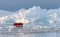 Antarctic expedition, cruise passengers in red parkas ride in a Zodiac inflatable boat, very close to a huge white iceberg in Cierva Cove bay