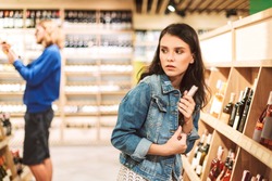 Young frightened woman in denim jacket trying steal bottle of wine in modern supermarket