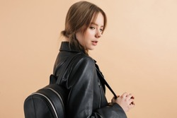 Portrait of young attractive girl with two braids in leather jacket with black backpack on shoulder standing from back dreamily looking aside over beige background