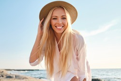 Young attractive smiling blond woman in white shirt and hat joyfully looking in camera with sea on background