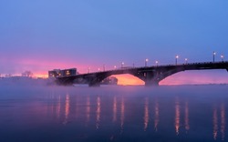 the bridge over the icy river in winter at sunset, at dawn. a snow-covered bank of a winter river at dawn, from which steam rises, and a bridge. Irkutsk, Siberia, Russia.