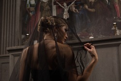 young blond woman with beautiful hairstyle is covered by a black veil in black dress kneeling at the altar in the background of paintings and arches and prays to Jesus looking away