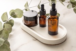 anti-aging collagen facial serum in dark glass bottle and face cream on white concrete tray on marble background with eucalyptus branch. Natural Organic Cosmetic Beauty Concept.