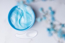 Eye patches in a plastic container, top view. Hydrogel patches with peptides for the area around the eyes. Blue gel patches with a plastic spatula in a jar on a marble background.