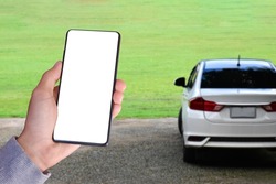 White blank touchscreen mobilephone holding in hand with blurred white car background. Close up view of humans hand with mobile device. Mock up, copyspace, template and technology concept.