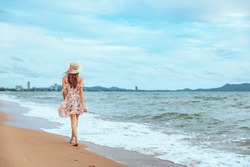Summer travel vacation concept, Happy traveler asian woman with dress and straw hat walking and open arm on sea beach at Pattaya, Thailand
