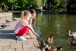Two cute little girls feeding  group of ducks in pond with seeds from their hands in a park on a sunny day. Children interact with birds. Children taking care of animals outdoors.