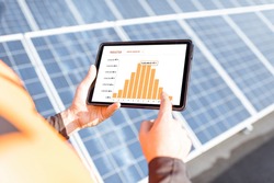 Workman examining genaration of solar power plant, holding digital tablet with a chart of electricity production. Concept of online monitoring of the electric station