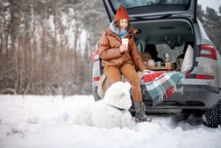 Young woman with her white dog have a picnic in car trunk near a pine forest, traveling by car during winter holidays. High quality photo
