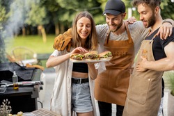Young people enjoy yummy burgers made on a grill at picnic, standing together and having fun. Friends cooking at backyard outdoors. American lifestyle