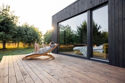 Young woman resting on sunbed and reading on a tablet on the wooden terrace near the modern house with panoramic windows near pine forest. Concept of solitude and recreation on nature