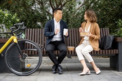 Asian businessman and caucasian businesswoman eating food and drinking coffee while having lunch at work. Concept of rest and break on job. Business people sitting on bench on city street