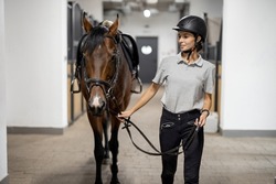 Female horseman going with her brown Thoroughbred horse in stable. Concept of animal care. Rural rest and leisure. Idea of green tourism. Young european woman wearing helmet and uniform