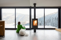 Woman sitting with dog near fireplace and panoramic window at modern living room with stunning view on snowy mountains. Concept of rest in houses or cabins on nature. Idea of escape from everyday life
