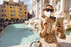 Woman in face mask with phonein front of famous di Trevi fountain in Rome. Concept of new social rules when traveling. Social distancing and wearing mask in public places