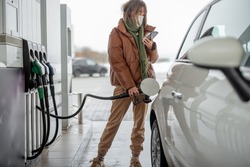 Woman in face mask refueling car with a gasoline, using smartphone to pay. Concept of mobile technology for fast refueling without visiting the store