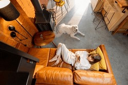 Woman lying relaxed on couch and using phone with her dog at home. Wide angle view from above. Spending leisure time with digital gadgets and pet