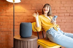 Young cheerful woman using smart gadgets at home, talking to smart speaker and listening to the music while sitting with phone on brick wall background