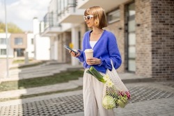 Woman using phone while standing with mesh bag full of fresh vegetables and reusable coffee cup on porch of her house. Concept of sustainability, healthy food and modern eco-friendly lifestyle