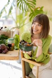 Young cheerful woman eats vegetarian lunch in bowl, sitting by the table full of fresh food ingredients indoors. Healthy lifestyle and wellness concept