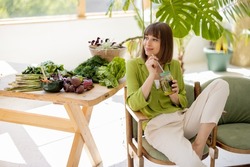 Young woman drinks lemonade while sitting on chair near table with lots of fresh food ingredients in room with green plants. Healthy lifestyle concept