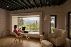 Woman works on laptop while sitting by the table in front of panoramic window with great view on mountains. Wide interior view. Remote work and escaping to nature concept
