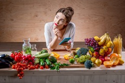 Young and pretty woman using smart phone sitting at the table full of fruits and vegetables in the wooden interior. Counting calories with mobile app. Food and health care concept
