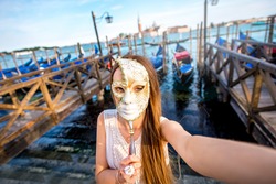 Young female traveler making selfie photo with carnaval mask standing near San Marco square with gondolas on the background in Venice.
