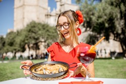 Young woman in red dress with sea Paella, traditional Valencian rice dish, sitting outdoors at the restaurant in the centre of the old town of Valencia