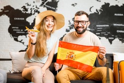 Young couple sitting with spanish flag at the travel agency office on the world map background prepairing for a trip to Spain
