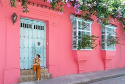 Side view of woman sit on a pink wall in Cartagena de Indias. Horizontal view of latin woman sightseeing in spanish historic ancient city. Travel to Colombia concept.