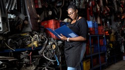 happy african american engineer warehouse worker checking list inventory of old used car engine in garage store. black woman  Inventory Manager in Recycle motor factory auto parts shop
